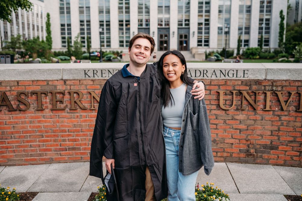 Two students stand together in front of a brick wall with Eastern University signage behind them on campus.