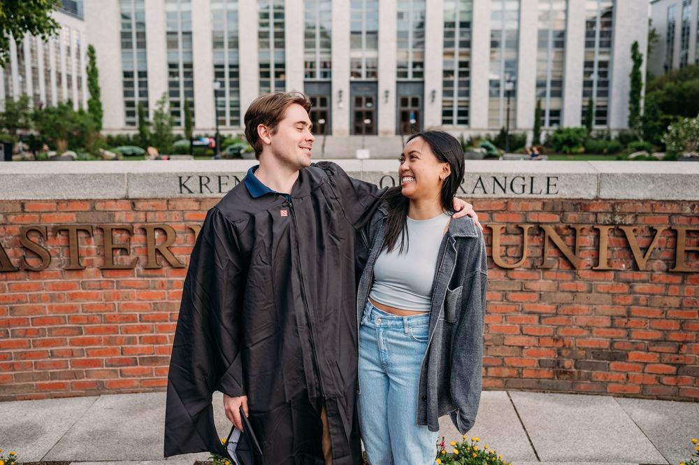 Two students in leather jackets smiling at each other in front of a brick university sign with a glass building behind them.