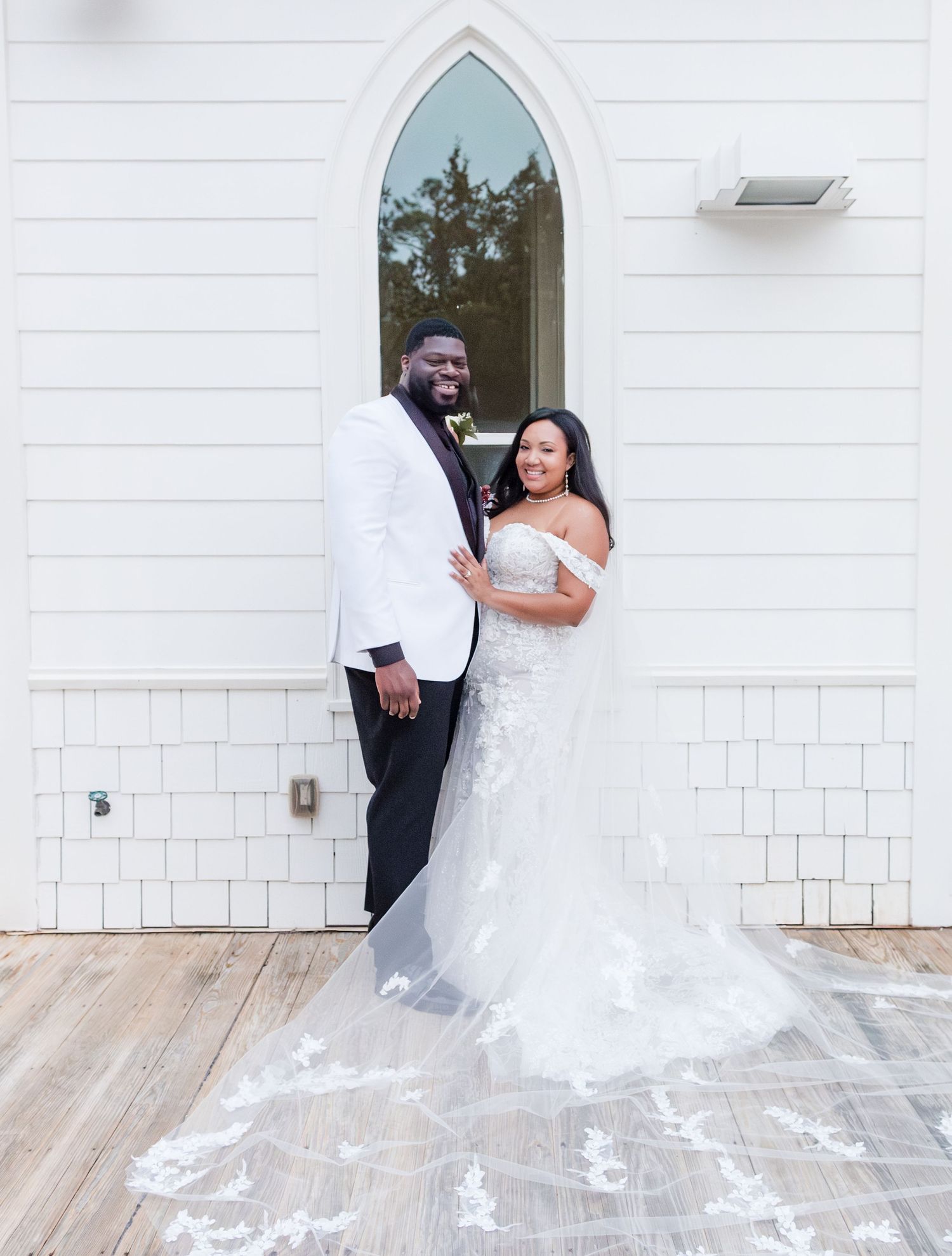 Elegant wedding couple poses in front of a white church with an arched gothic window and shiplap siding.