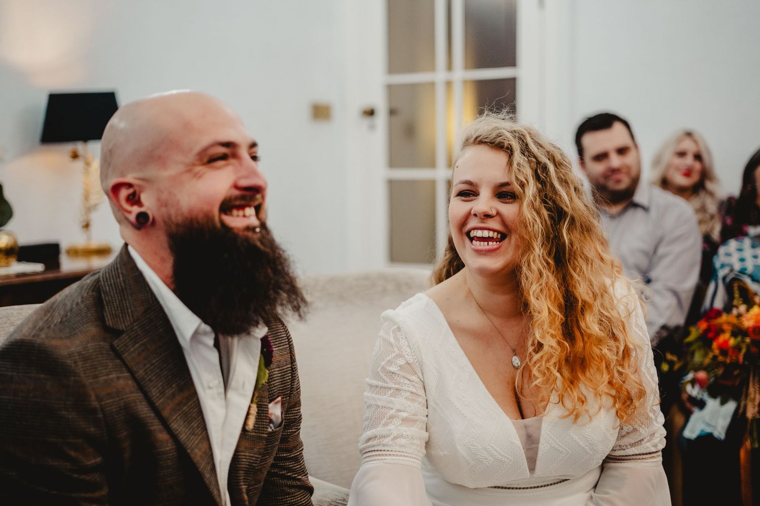 A joyful couple in wedding attire share a candid laughing moment while guests smile in the background.