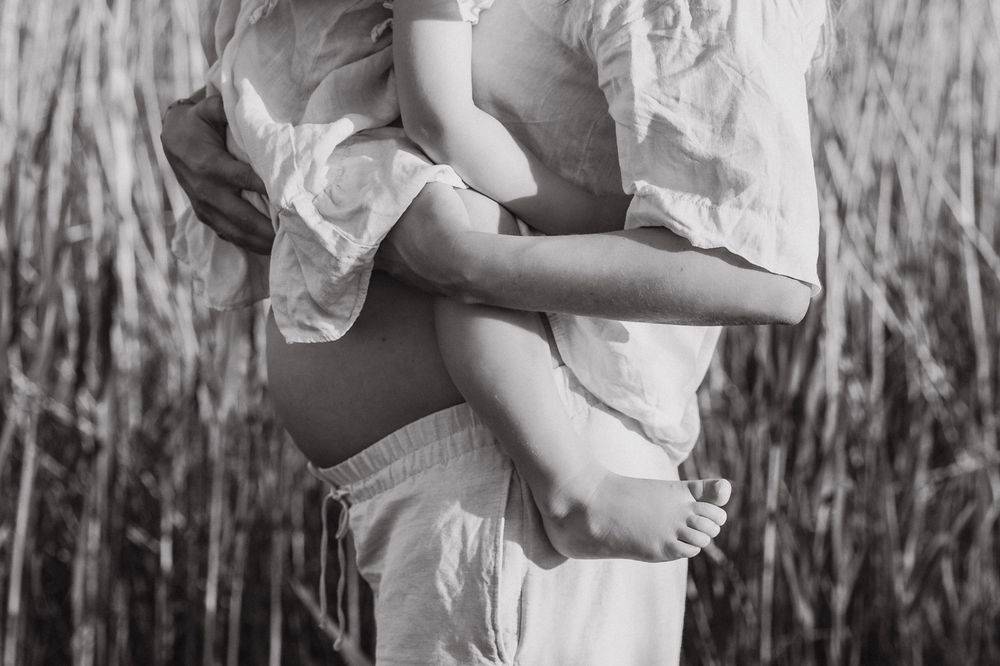 Black and white maternity photo showing pregnant belly and delicate white dress against tall reeds in nature.