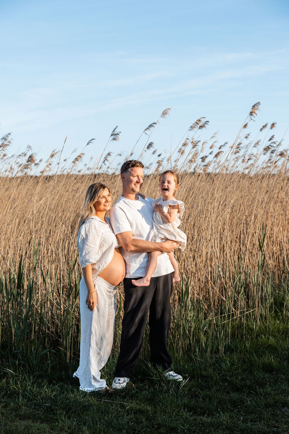 A couple standing in a tall grass field on a sunny day wearing white casual clothing.