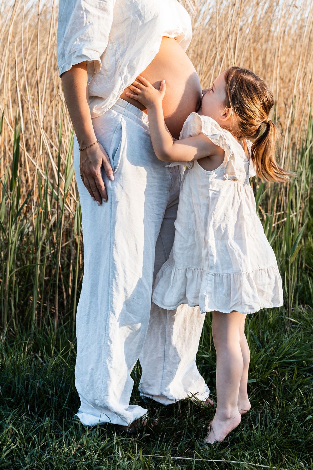 A young child in a white dress embraces an adult in white clothing while standing in a field of tall reeds at sunset.