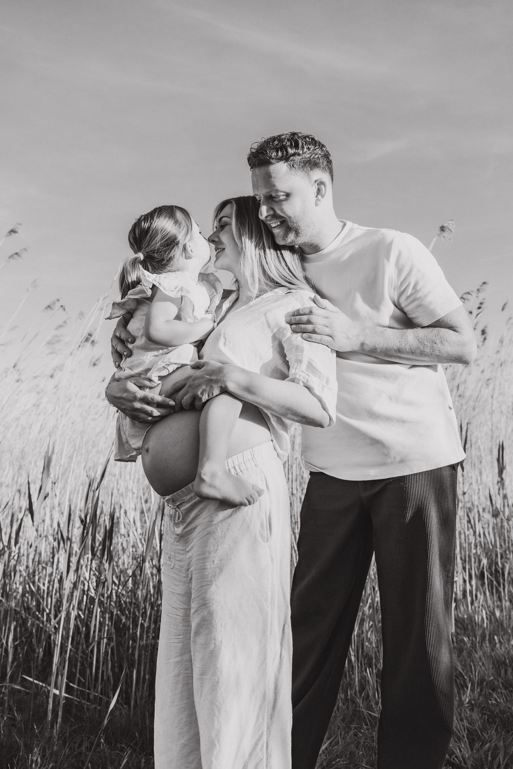 Black and white outdoor portrait of family sharing intimate moment together in tall grass field under cloudy sky.