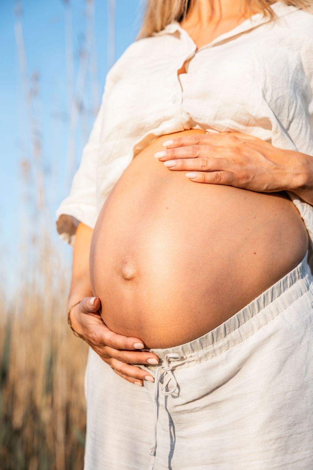 Woman in white clothing tenderly holds her pregnant belly against a natural outdoor background at sunset.