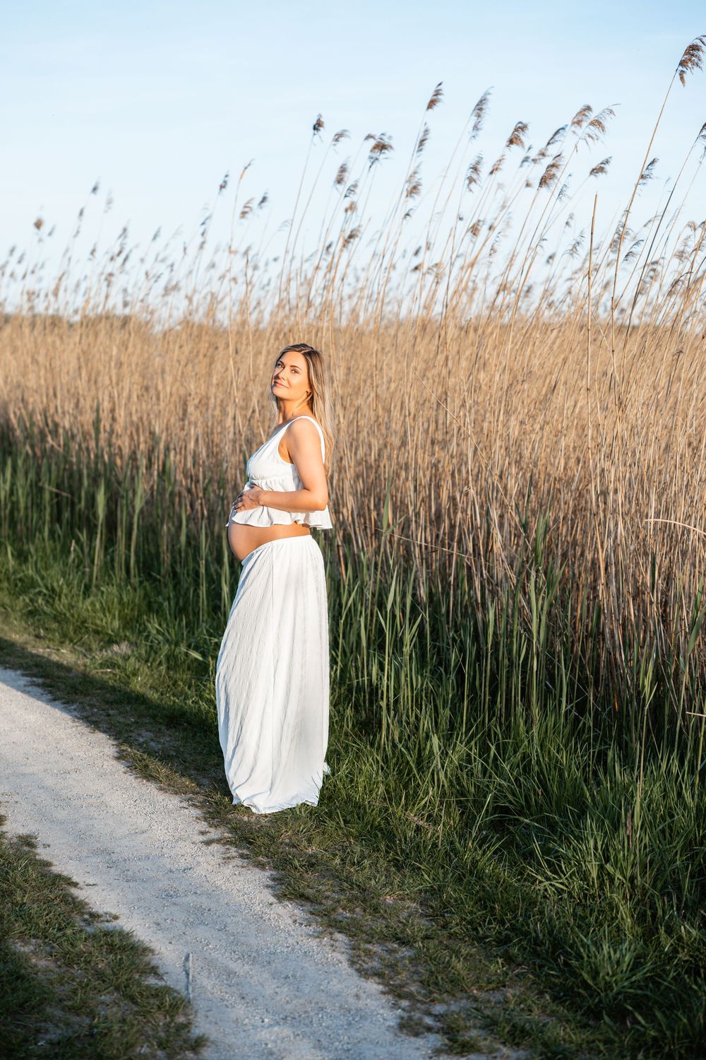 A pregnant woman in a flowing white dress stands on a path beside tall reeds at golden hour.