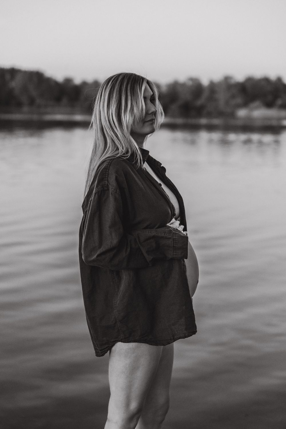 Black and white portrait of a pregnant person wearing an open button-down shirt while standing near a calm lake.