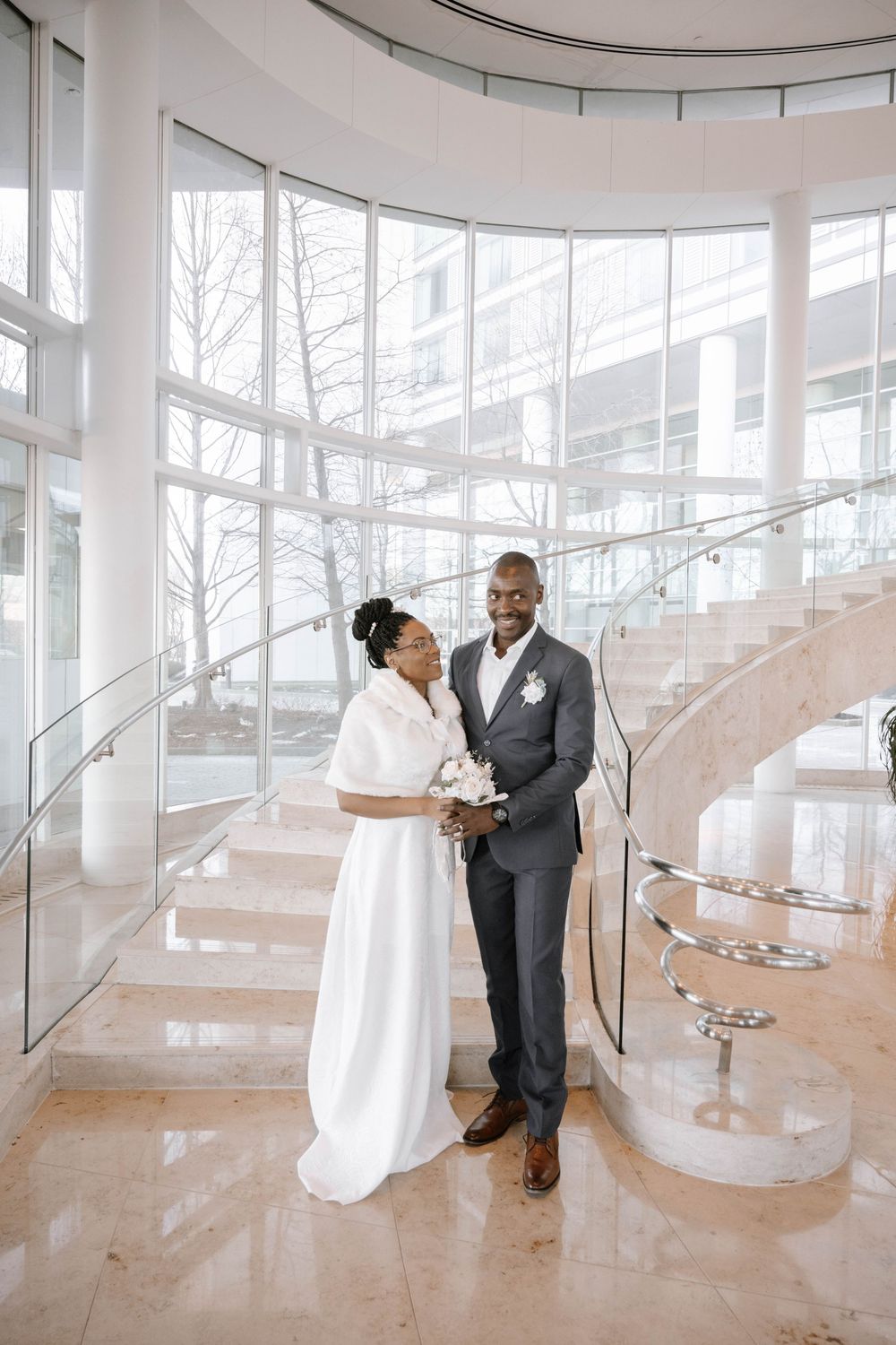A wedding couple poses elegantly in front of a curved glass staircase in a modern bright building interior.