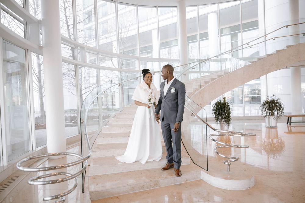 Elegant couple stands in a modern atrium with curved glass staircase and natural light streaming through windows.