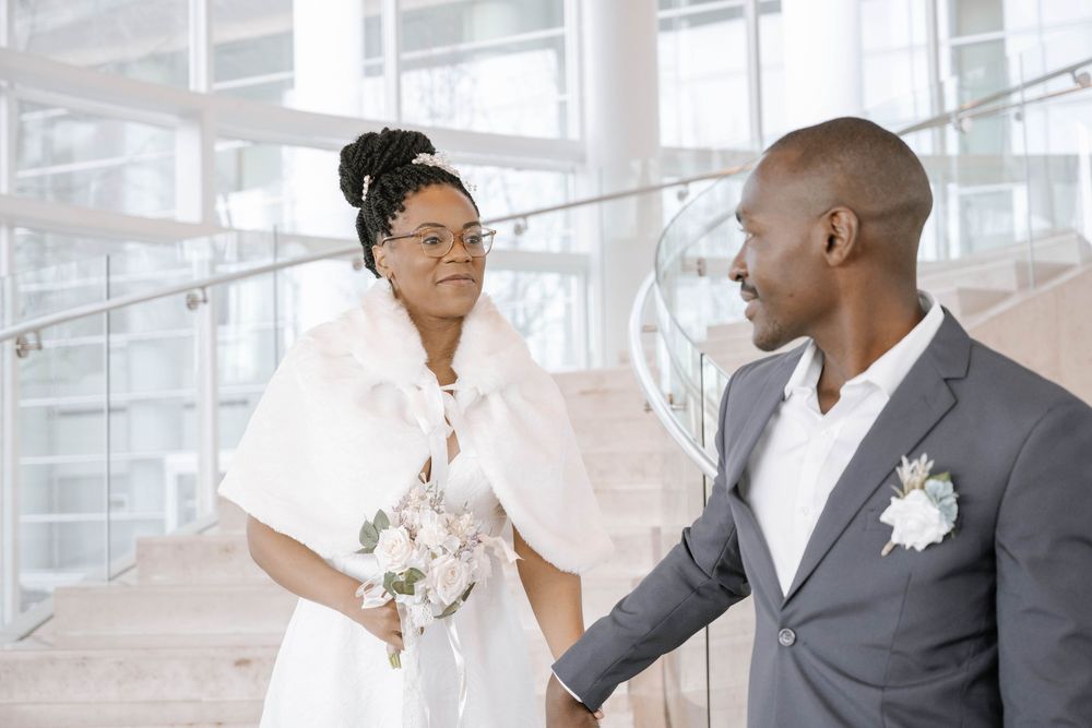 Elegant wedding couple in white gown and grey suit sharing a tender moment in a sun-lit modern building.