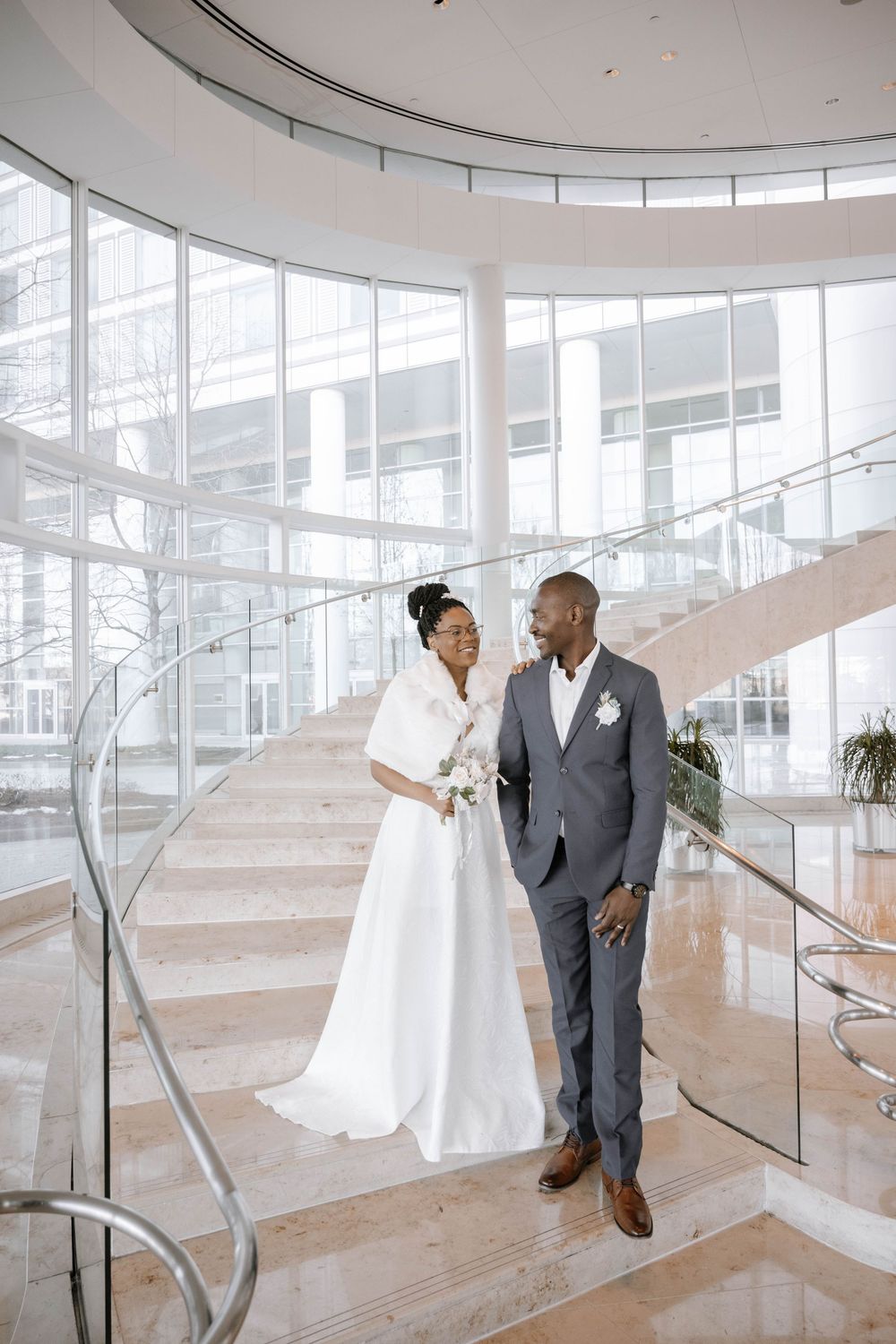 A couple in wedding attire stands elegantly on curved white marble stairs in a modern building.