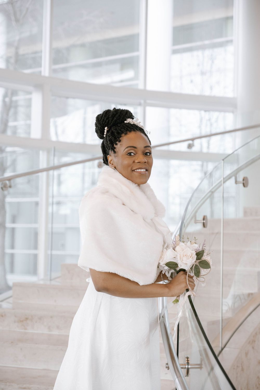 Bride wearing a white wedding dress and plush fur shawl holds pink bouquet while standing on modern staircase.