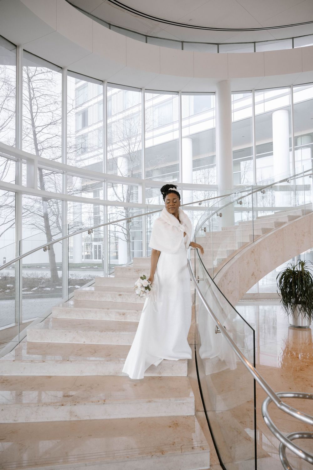 Person in a flowing white dress poses on modern marble staircase with curved glass walls and natural lighting.