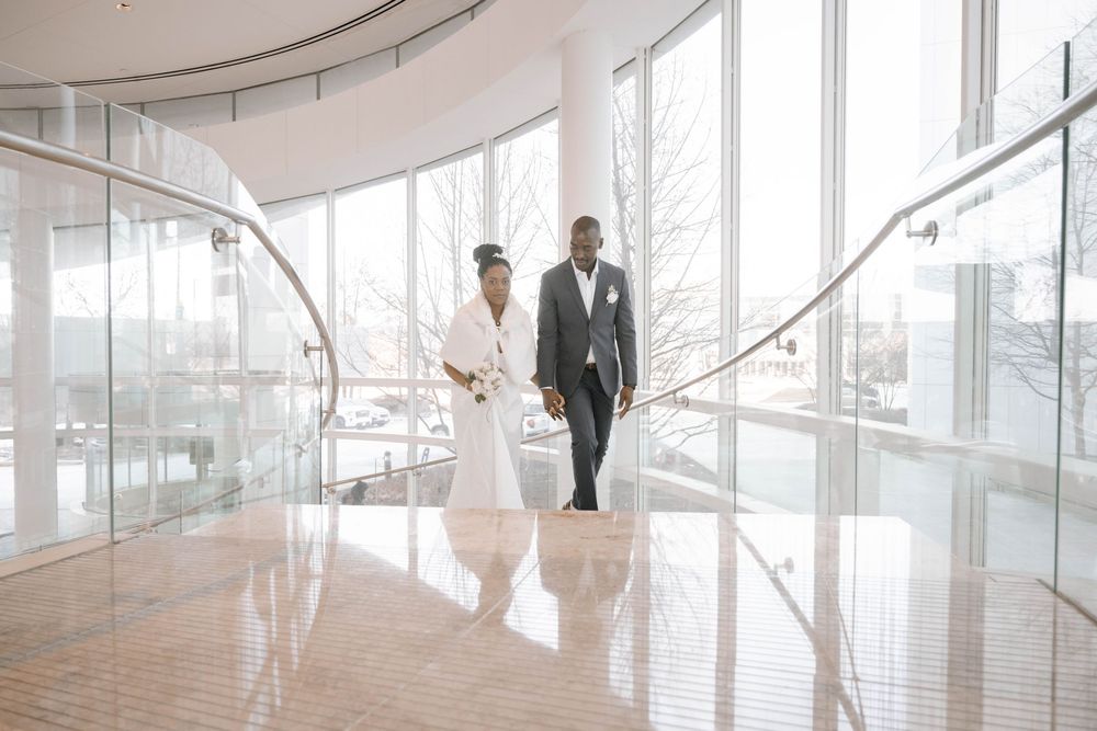 Two people in formal attire walk through a modern glass-lined corridor with natural light streaming in.