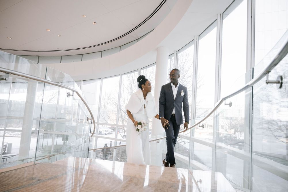 A couple in wedding attire walking through a modern curved glass building with winter views outside.