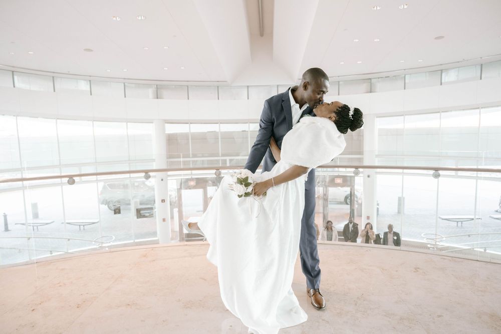 A romantic wedding couple shares a dip-style dance pose in a bright circular atrium with large windows.