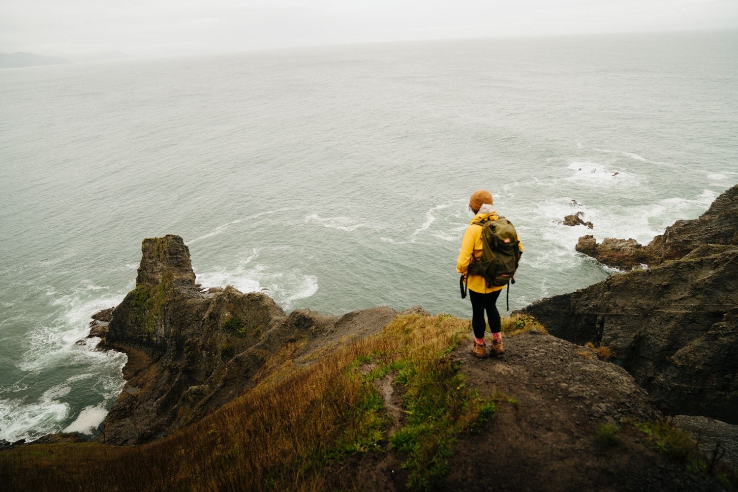 A person in a yellow raincoat stands on a rocky coastal cliff overlooking stormy ocean waves.