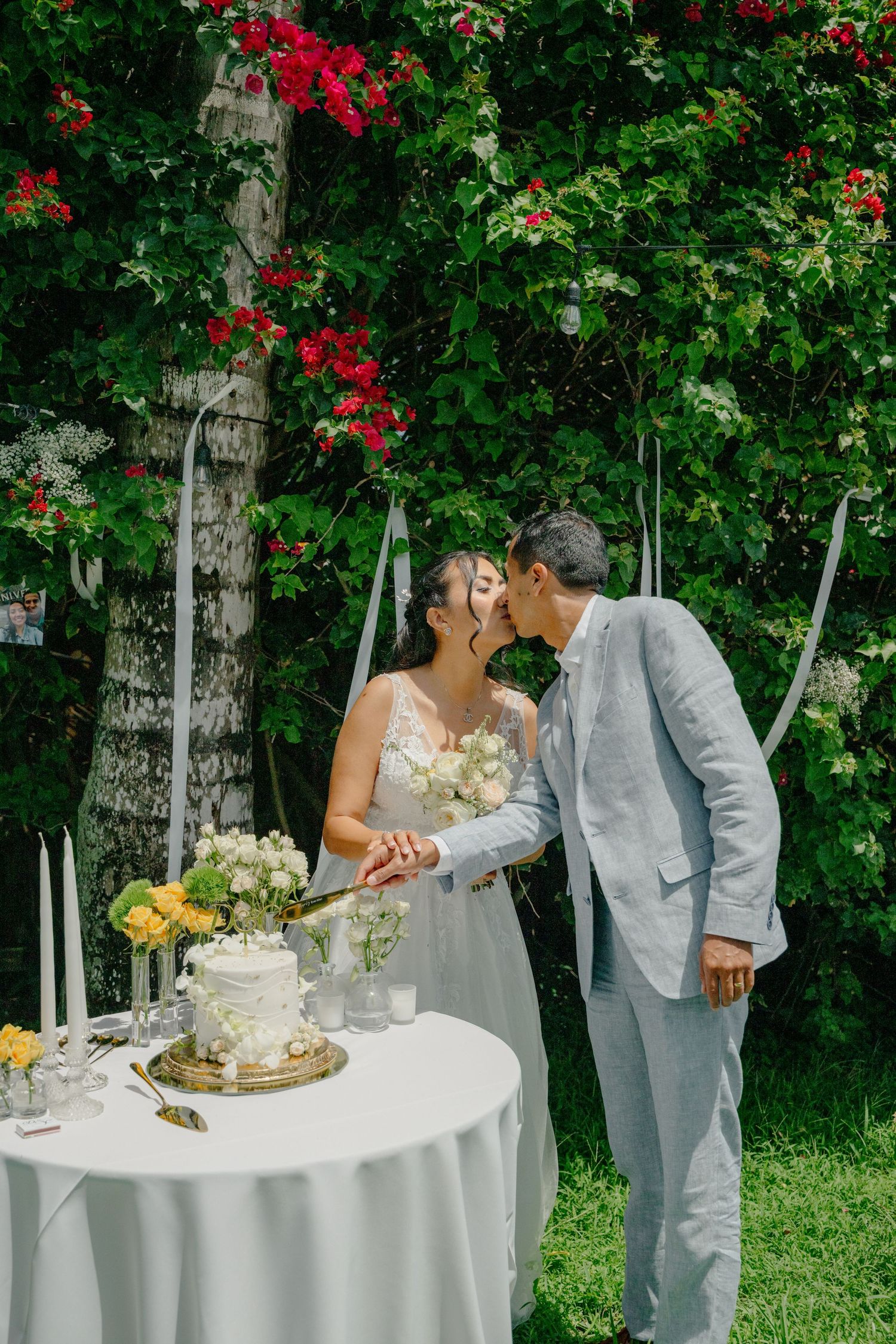 A romantic wedding couple shares a kiss next to an elegantly decorated cake table with red bougainvillea flowers in the background.
