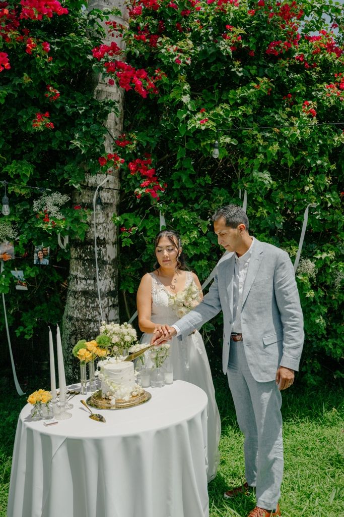 A romantic garden wedding scene with a couple cutting their cake at a white-clothed table beneath blooming red bougainvillea.
