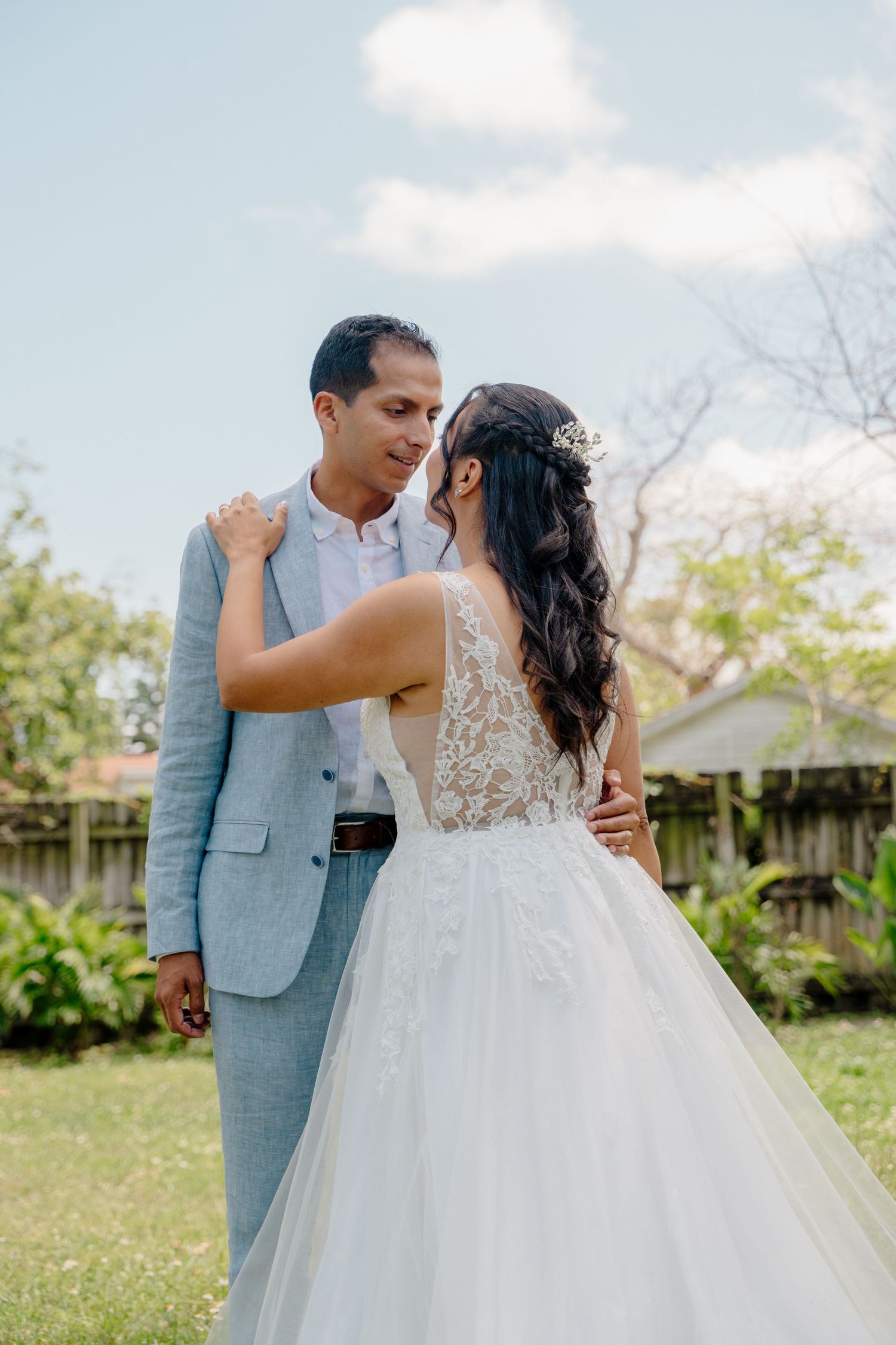 A couple embracing in a garden setting during their wedding, the bride in a lace white gown and groom in a light gray suit.
