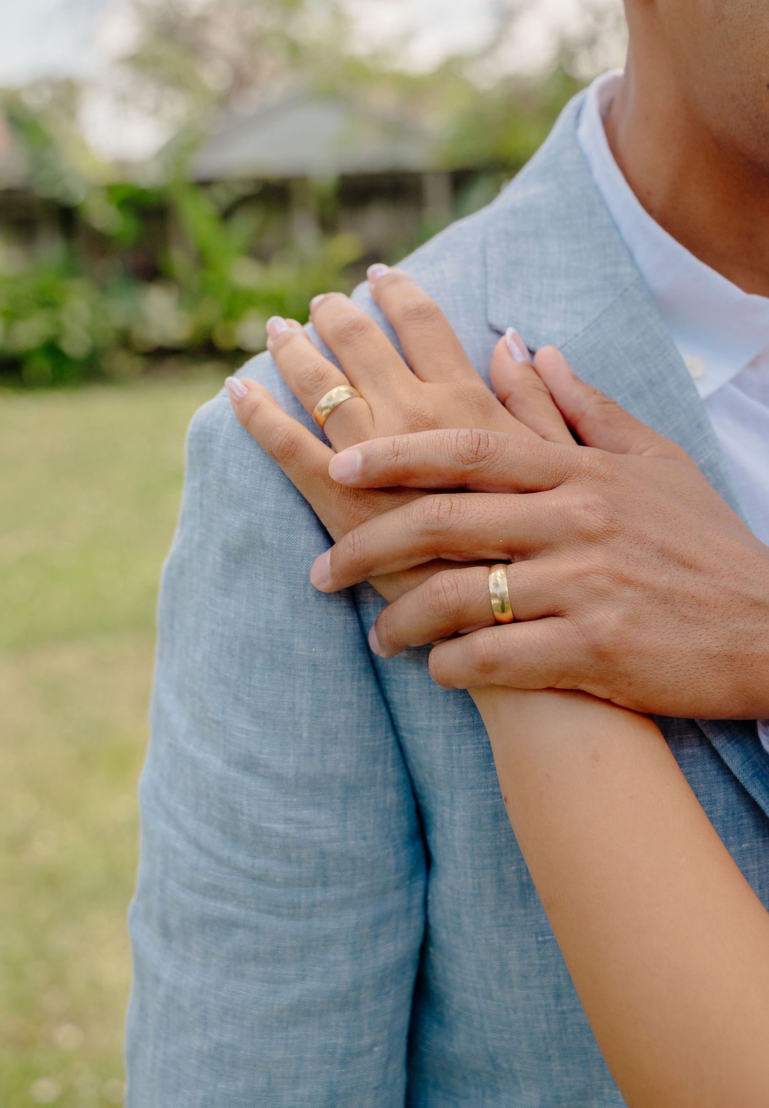 Two hands with wedding rings embrace tenderly against a light blue suit jacket in an outdoor garden setting.