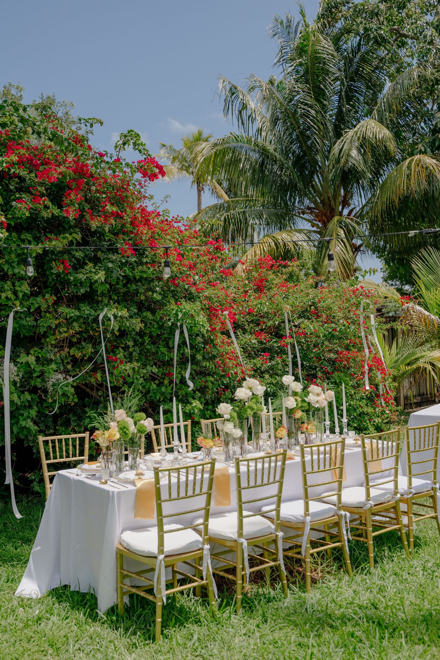 Elegant outdoor wedding table setup with gold Chiavari chairs and floral centerpieces surrounded by tropical palm trees and bougainvillea.