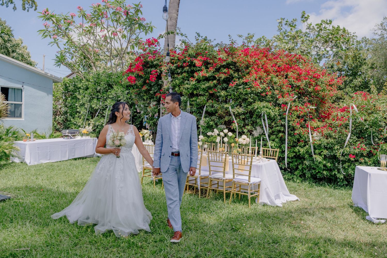 Outdoor garden wedding ceremony setup with vibrant red flowers and white chairs on a sunny day.