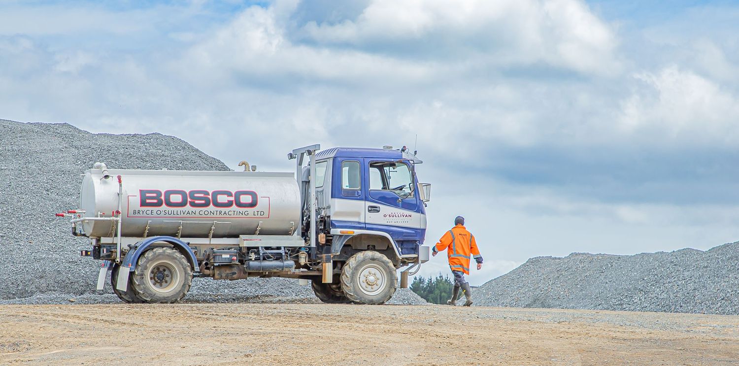Oil delivery tanker truck parked on dirt road with a worker in orange safety gear standing beside it in mountainous terrain.