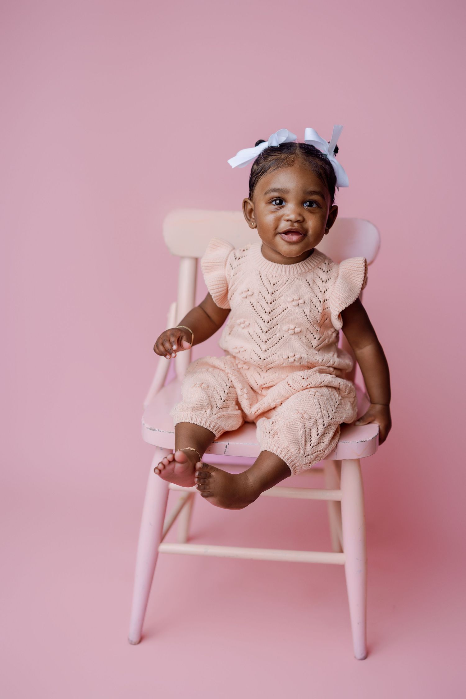 A happy toddler sits on a pink wooden chair wearing a light pink lace dress with ruffled sleeves against a pink backdrop.