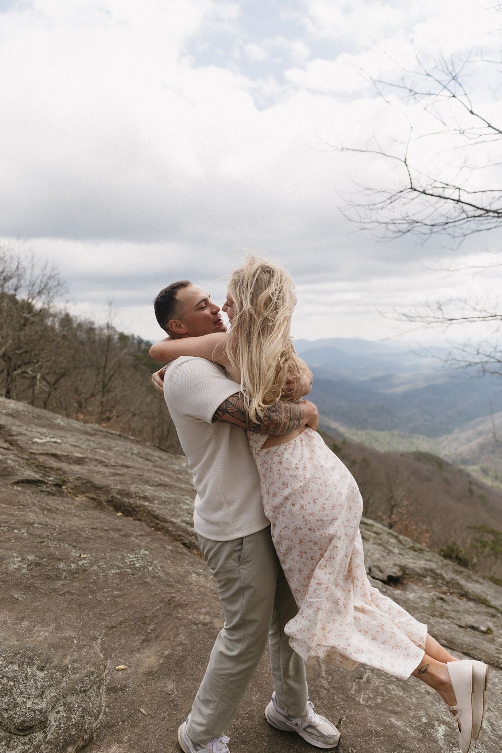 A couple embraces joyfully on a blue ridge mountain overlook with sweeping valley views and cloudy skies in the background.
