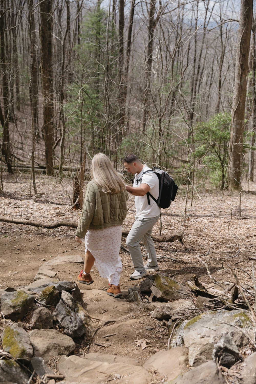 a couple stands together on a rocky hiking trail surrounded by bare trees and scattered evergreens on the appalachian trail