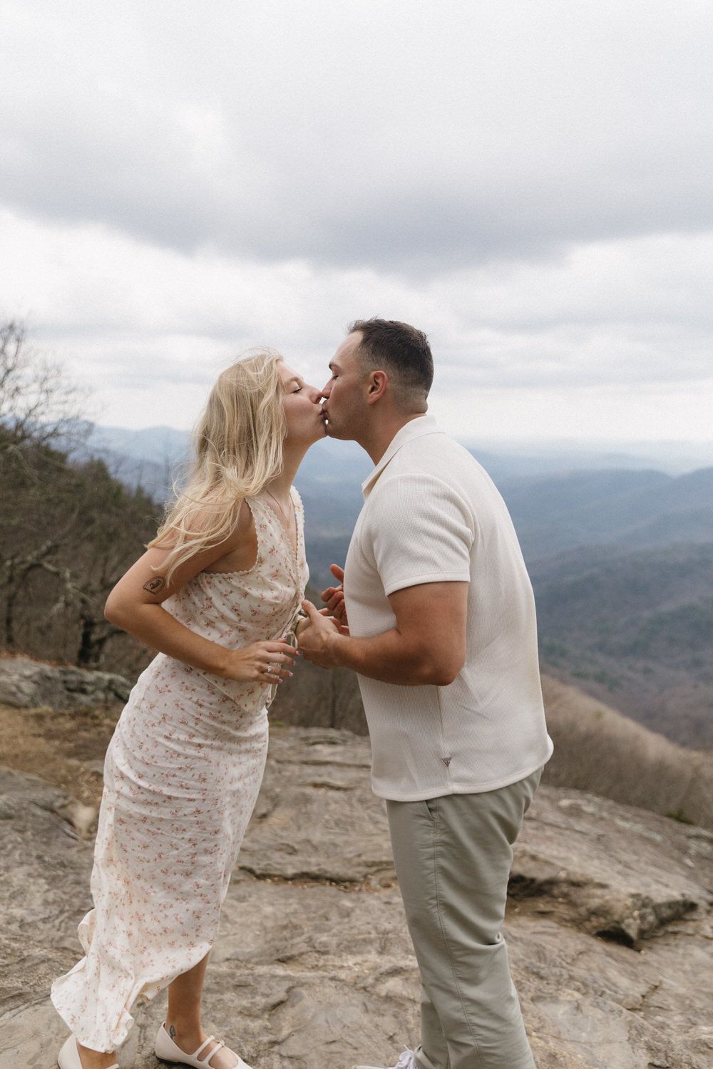 A couple shares their first kiss on a mountain overlook with misty blue ridges stretching into the cloudy horizon in north georgia