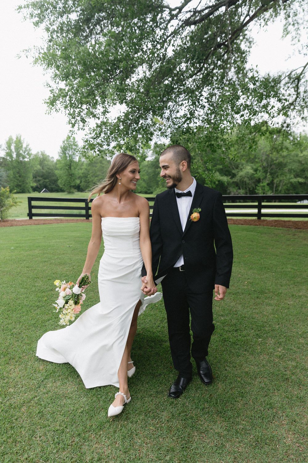 A couple walks together on a grassy lawn with a wooden fence behind them, sharing a romantic moment at their elopement