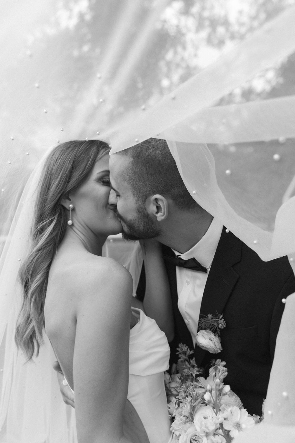 A romantic black and white wedding photo shows a couple sharing a kiss under a flowing veil against sunlit clouds.