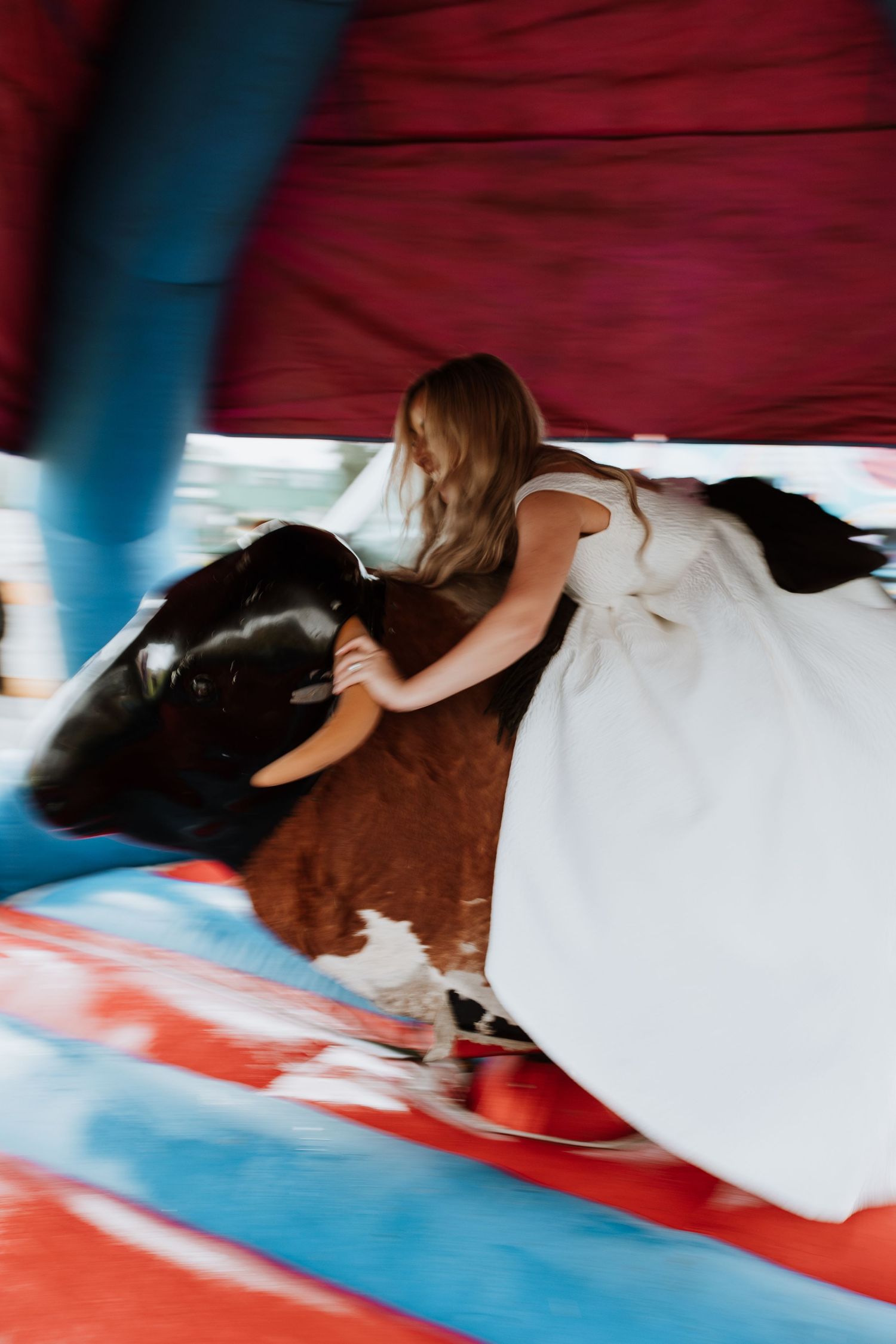 Bride riding a mechanical bull at a carnival with red and blue lighting and background decor.