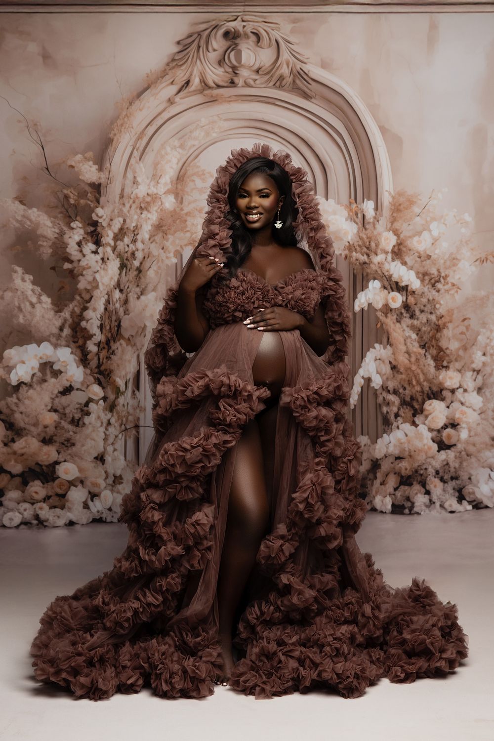 A stunning maternity photo featuring a brown ruffled gown in front of an ornate arch surrounded by dried floral arrangements.