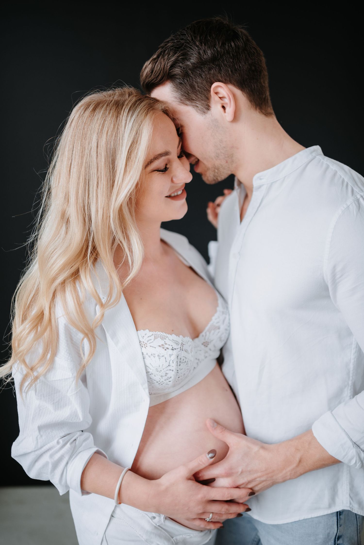 Intimate maternity photo featuring a couple in white clothing sharing a tender moment against a dark background.
