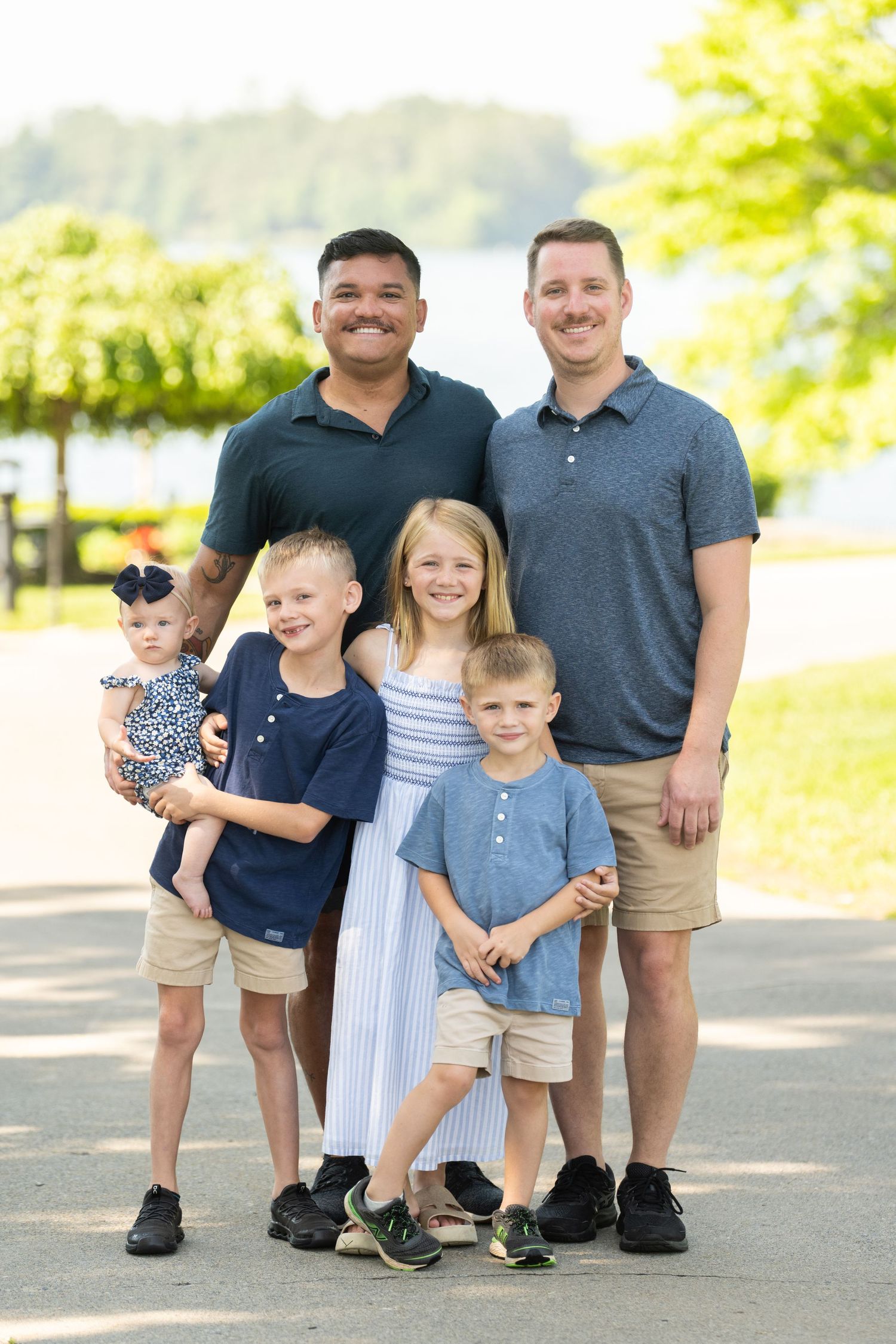 Happy family of six people standing together outdoors on a sunny day with trees in the background.