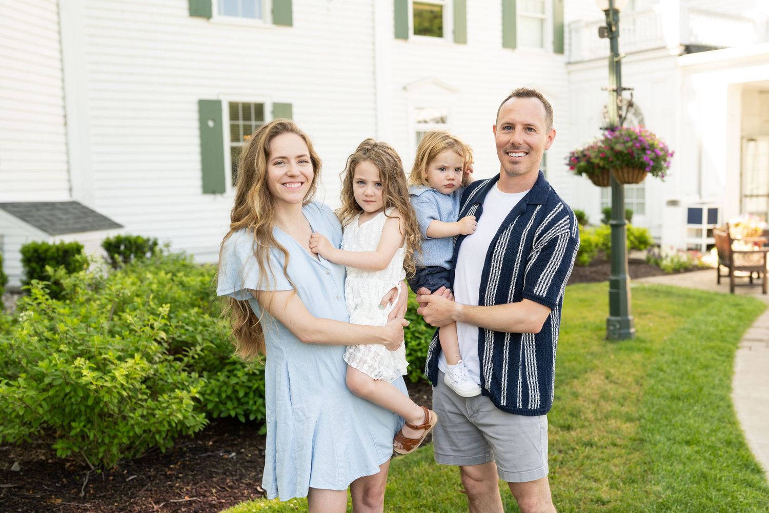 A family stands smiling together on a landscaped lawn outside a white house on a sunny day.