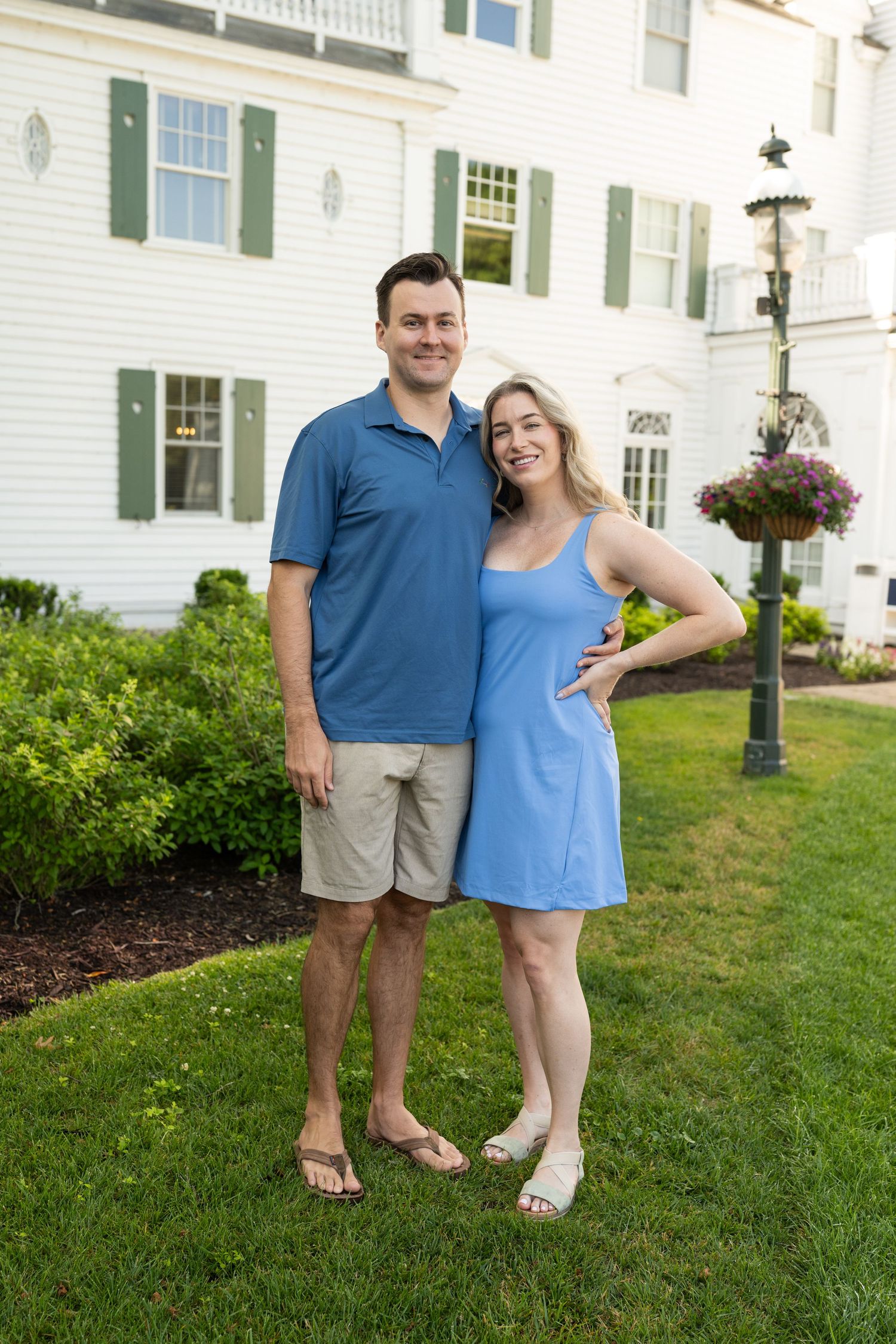 A couple in coordinating blue outfits stands on a manicured lawn in front of a white colonial-style house with green shutters.