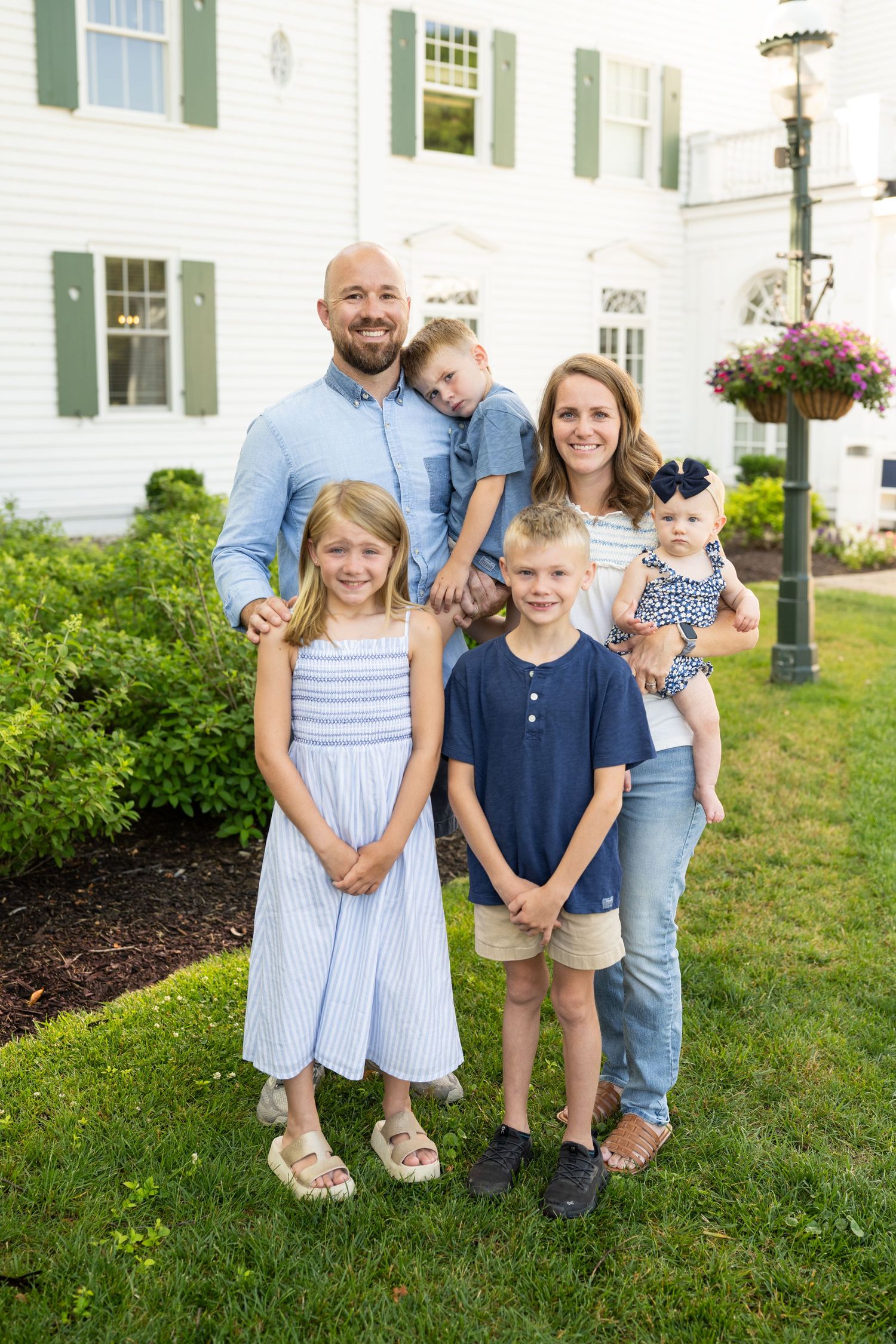 A group poses together on a lawn in front of a white house with green shutters and flowering landscaping.
