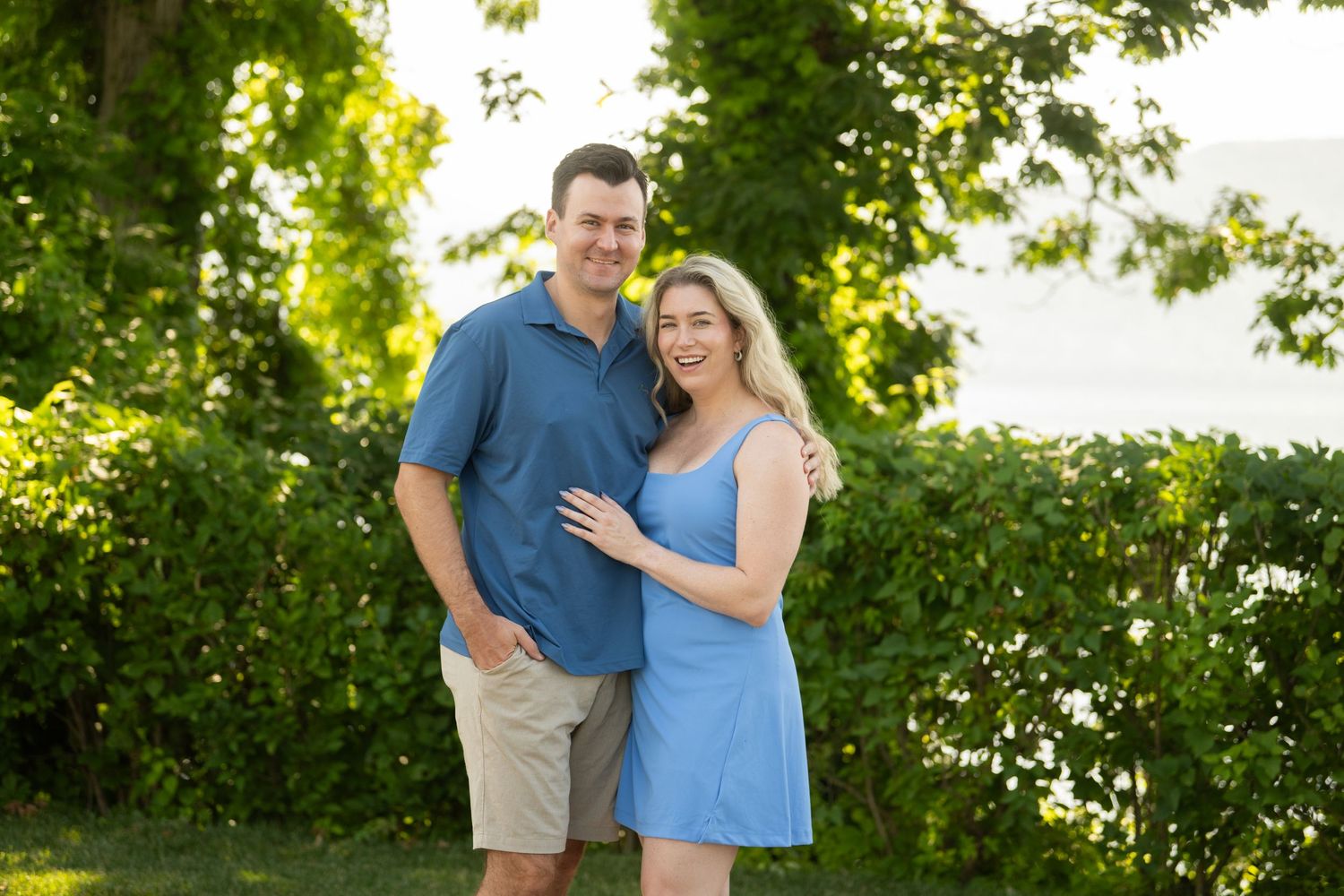 A couple poses together outdoors in a garden setting with lush green foliage in the background.