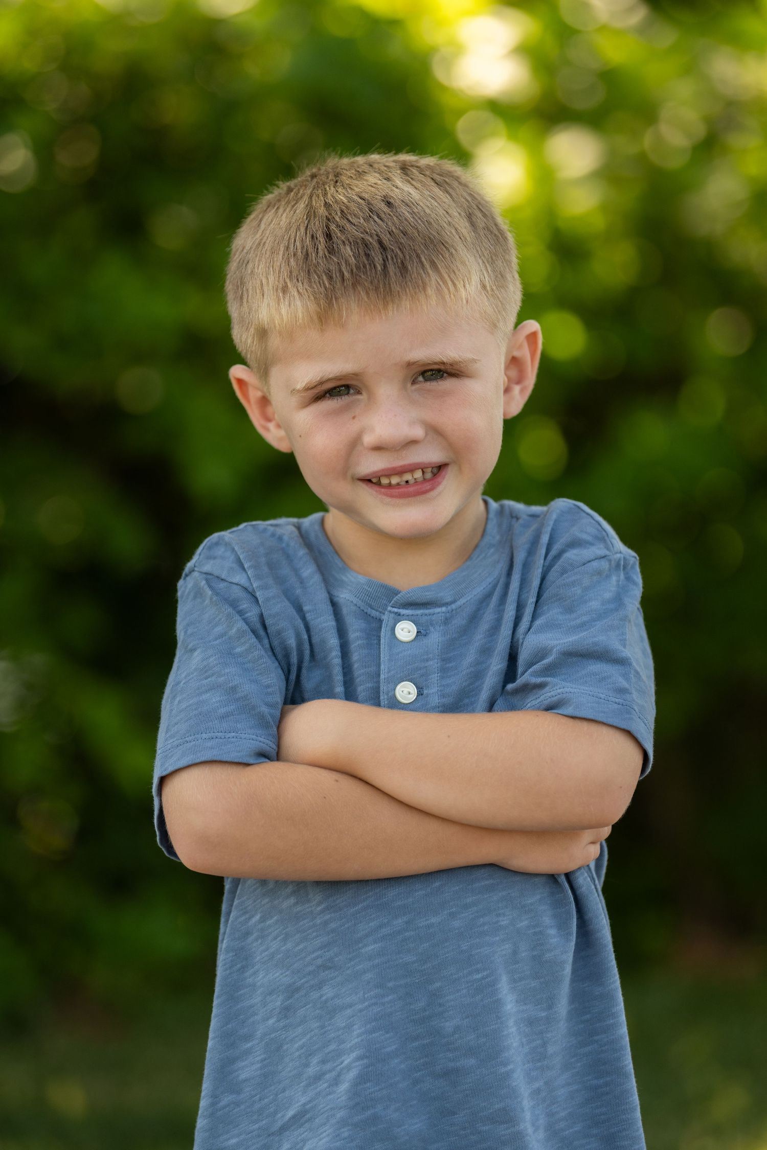 A young child in a blue henley shirt stands outdoors with arms crossed and a cheerful expression.