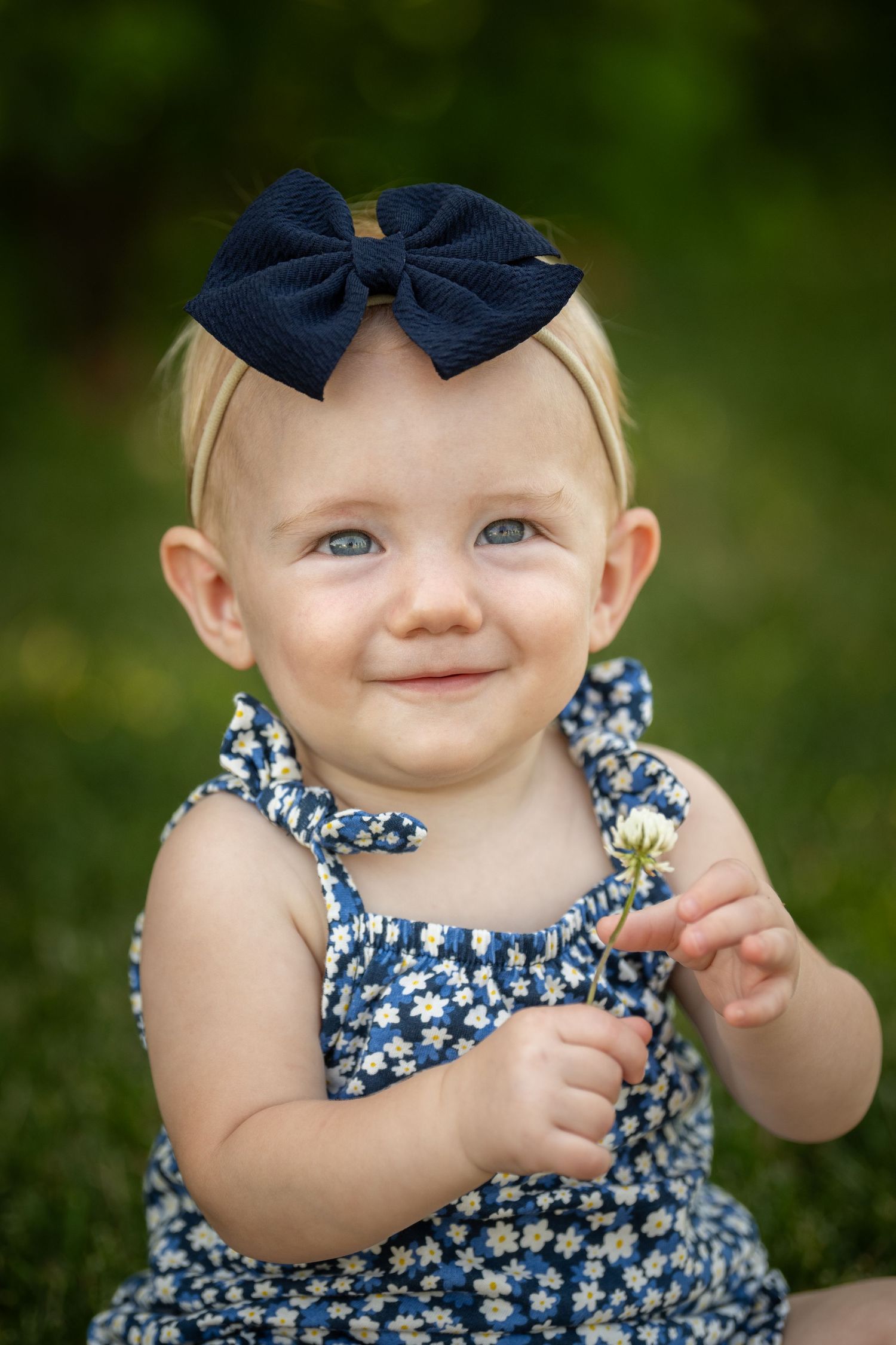 Smiling toddler in blue floral dress with navy bow headband holds small wildflower outdoors.
