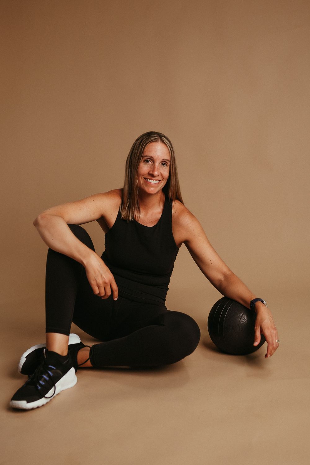 Athletic person in black workout attire sits on floor with a medicine ball against brown backdrop.