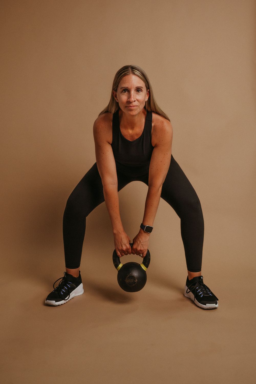 Person in black athletic wear performs a kettlebell workout against a brown backdrop.