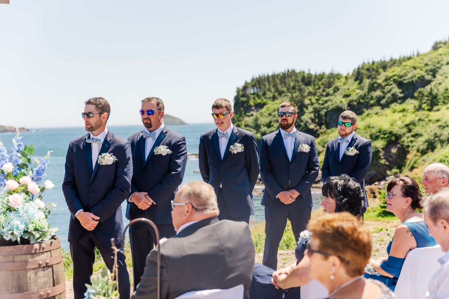 A wedding ceremony takes place outdoors on a sunny day with groomsmen standing in navy suits by the oceanside.