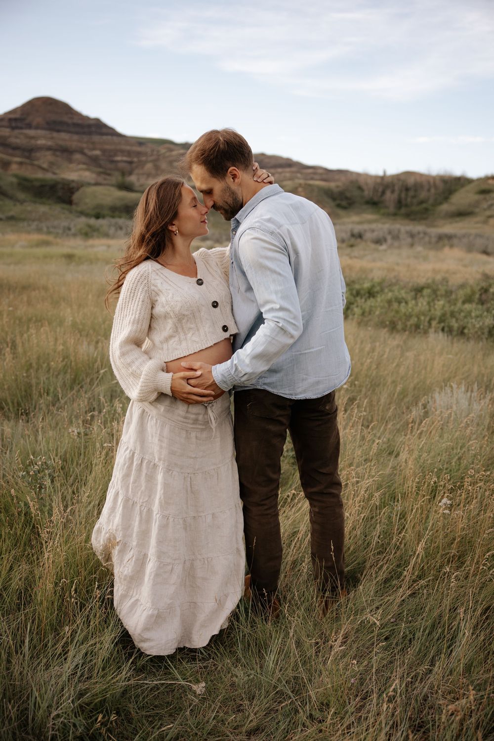 A romantic couple embraces in tall grass with rugged mountains in the background during golden hour.