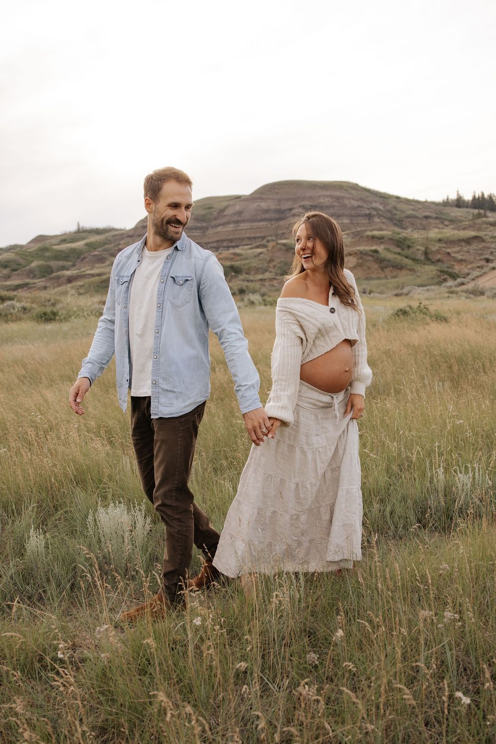A couple in casual attire walks and embraces through a rustic mountain meadow with tall grasses and rolling hills at sunset.