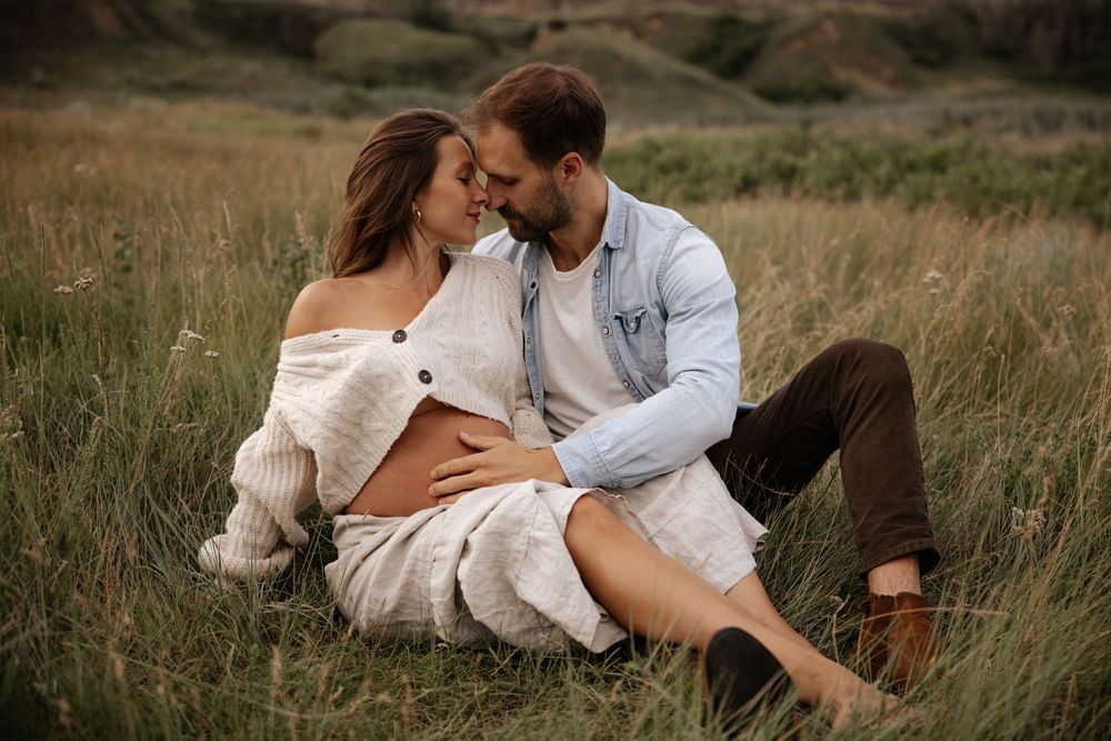 A romantic couple sits intimately together in a grassy field during golden hour.